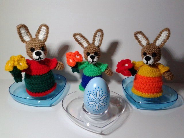 Drei gehäkelte Osterhasen-Eierwärmer in bunten Outfits mit Häkelblumen und einem blauen Osterei auf transparenten herzförmigen Schalen.