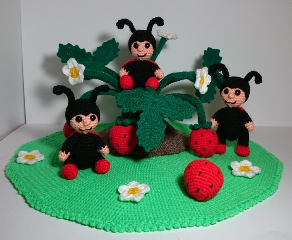 Drei gehäkelte Glückskäfer um eine Erdbeerpflanze mit roten Erdbeeren und weißen Blüten auf rundem, grünem Häkeluntergrund.