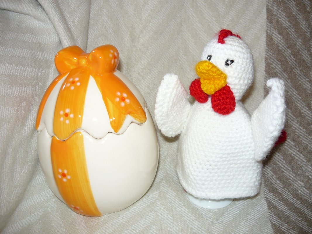 Weißer gehäkelter Huhn-Eierwärmer mit gelbem Schnabel und rotem Kamm neben einer orange verzierten Keramik-Eierschale