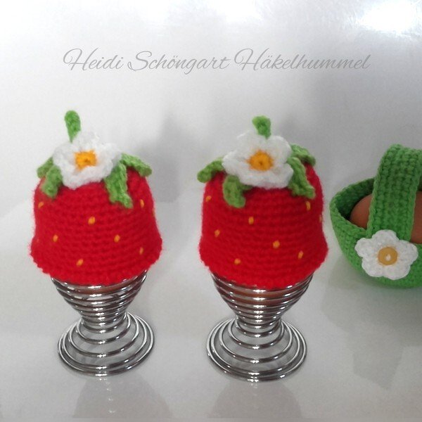Zwei rote, gehäkelte Erdbeer-Eierwärmer mit grünen Blättern und weißen Blüten auf spiralförmigen Metall-Eierhaltern; rechts ein kleiner grüner Häkelkorb mit Eiern.