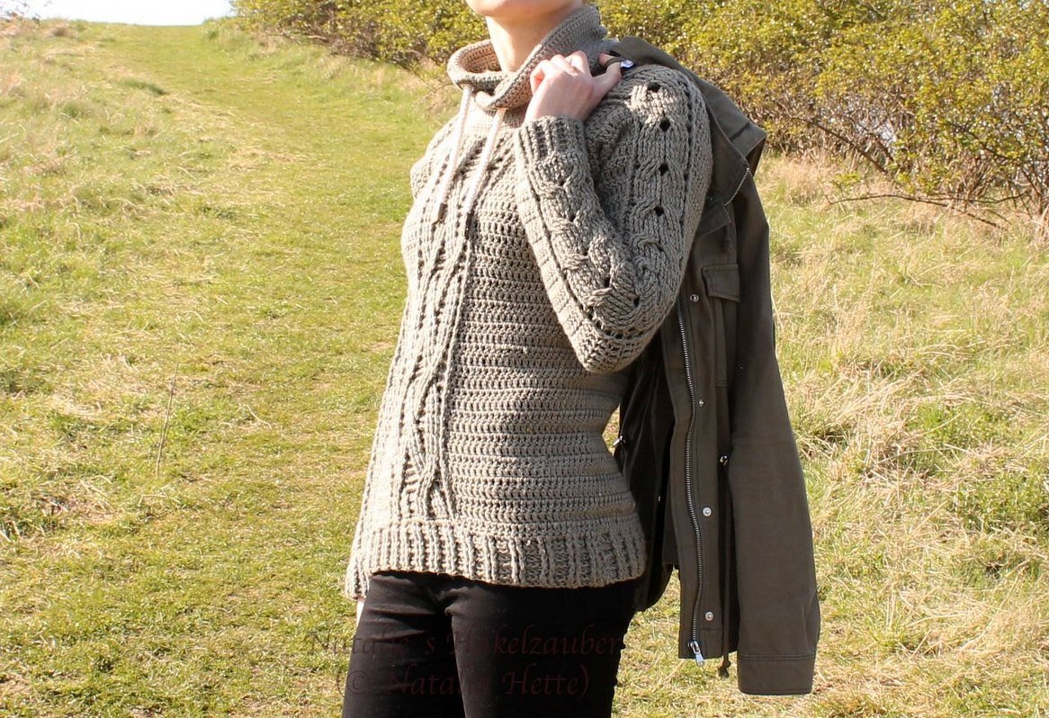 Woman wearing a greige crocheted hooded sweater with cable panels and openwork sleeves, standing on a grassy hill.