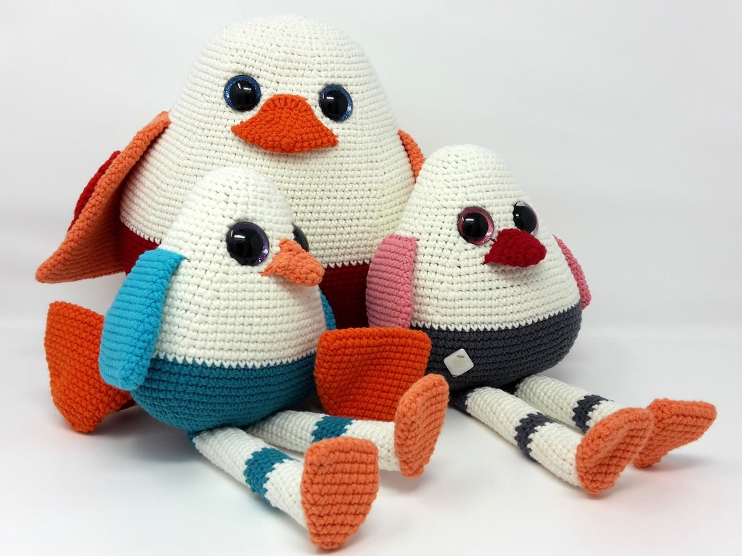 Drei gehäkelte Amigurumi-Vögel in Weiß mit orangen Schnäbeln, bunten Flügeln und gestreiften Beinen