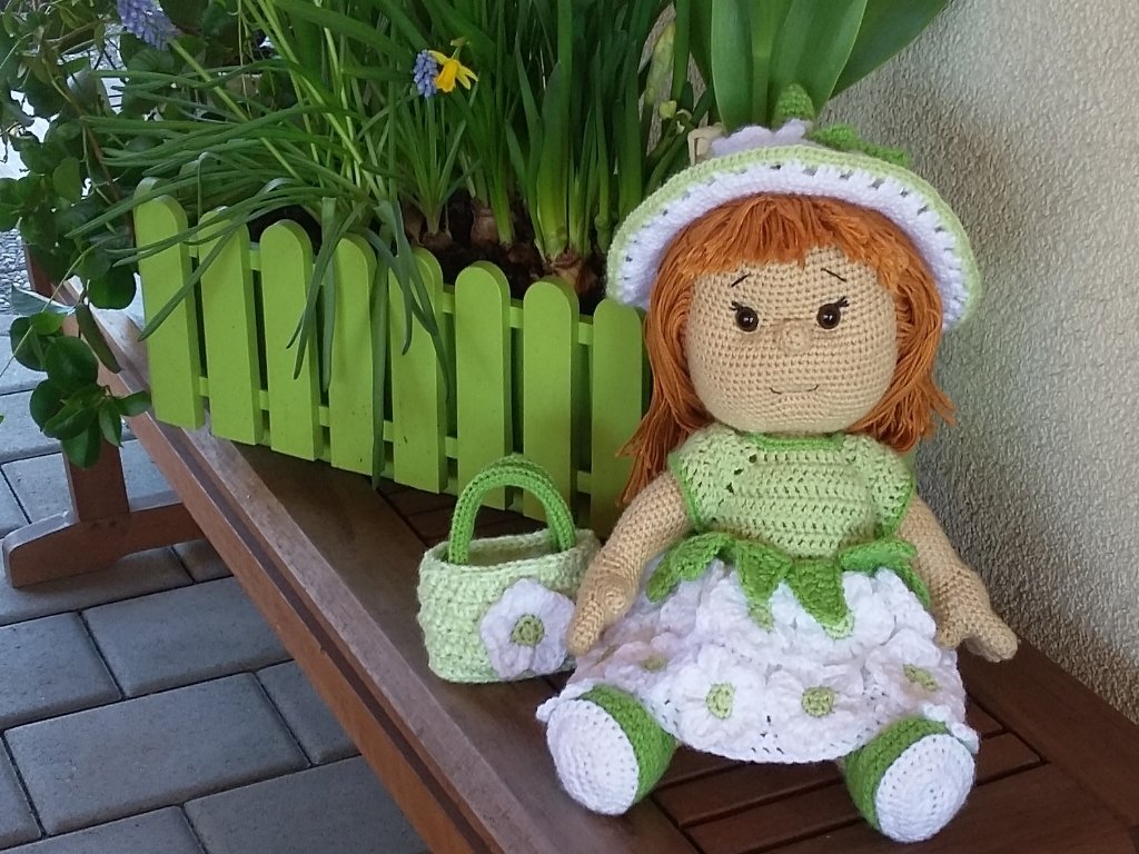 Gehäkelte Amigurumi-Puppe mit roten Haaren, weiß-grünem Blumenkleid und Sonnenhut neben kleinem gehäkelten Korb auf einer Holzbank vor einem grünen Blumenkasten.