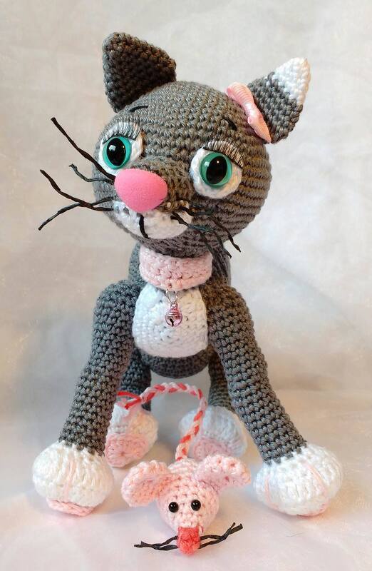 Gehäkelte graue Amigurumi-Katze mit rosa Nase, grünen Augen, weißen Pfoten und rosa Halsband, davor eine kleine rosa Häkelmaus an Kordel.
