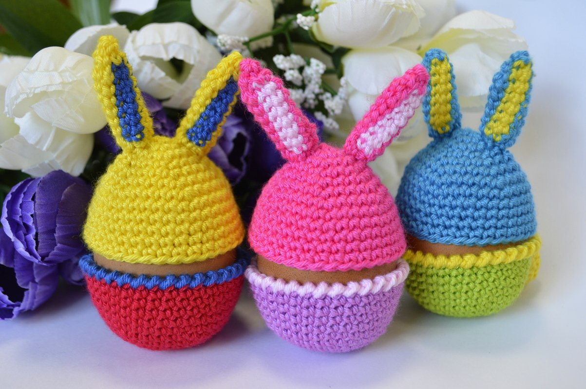 Drei bunte gehäkelte Eierwärmer mit Hasenohren vor Frühlingsblumen