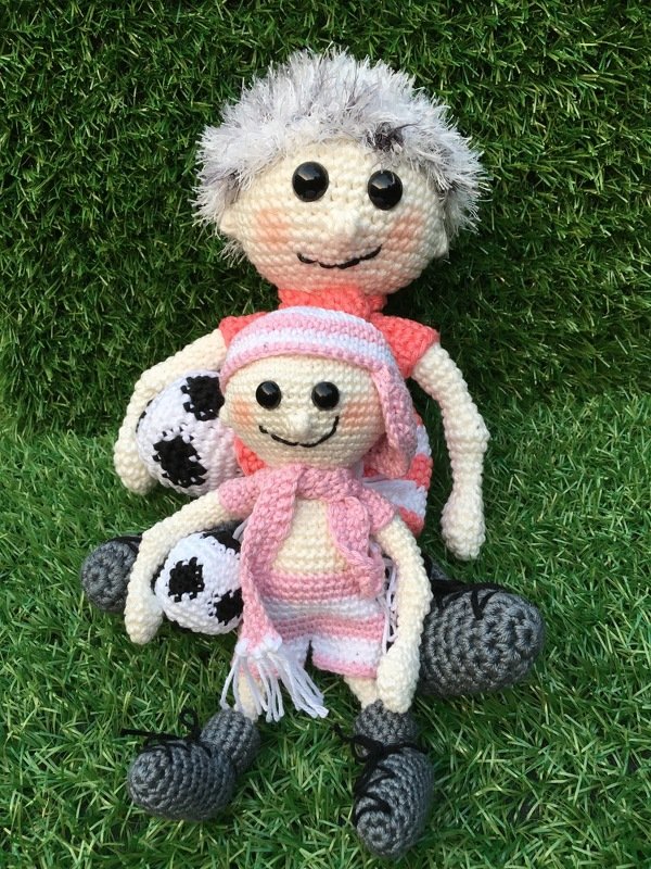 Zwei Amigurumi-Puppen (groß und klein) mit Fanbekleidung und je einem Fußball auf grünem Rasen.