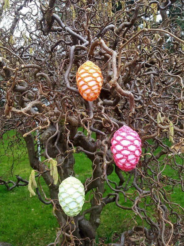 Drei mit Häkelnetz umhüllte Ostereier in Orange, Pink und Hellgrün, hängend an einem Baum mit gewundenen Ästen.