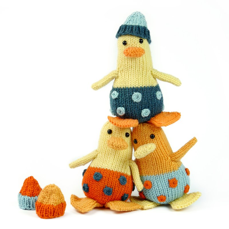 Three knitted amigurumi ducks stacked in a pyramid with two small knitted hats beside them.