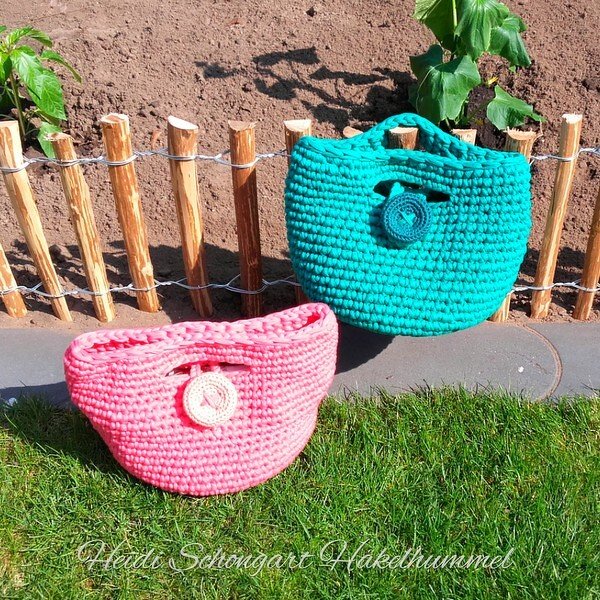 Zwei gehäkelte Korbtaschen in Pink und Türkis mit Knopf, vor einem Holzzaun auf Rasen