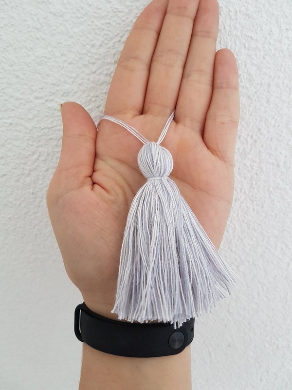 Hand hält hellgraue Garnquaste mit rund gebundenem Kopf und Aufhängeschlaufe vor heller Wand