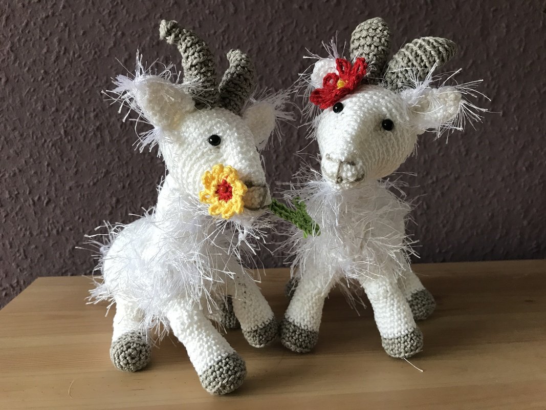 Zwei gehäkelte weiße Ziegen-Amigurumi mit grauen Hörnern und gehäkelten Blumen auf Holz