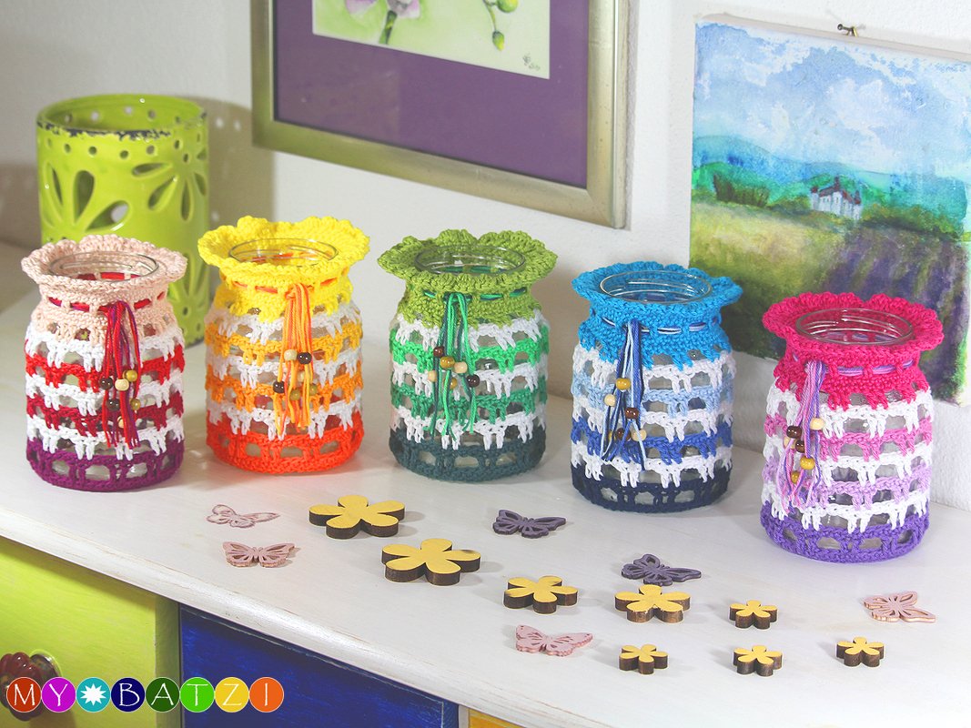Fünf Glasgefäße mit gehäkelten Hüllen in Rot, Orange, Grün, Blau und Pink, dekoriert mit Holzschmetterlingen und Blüten