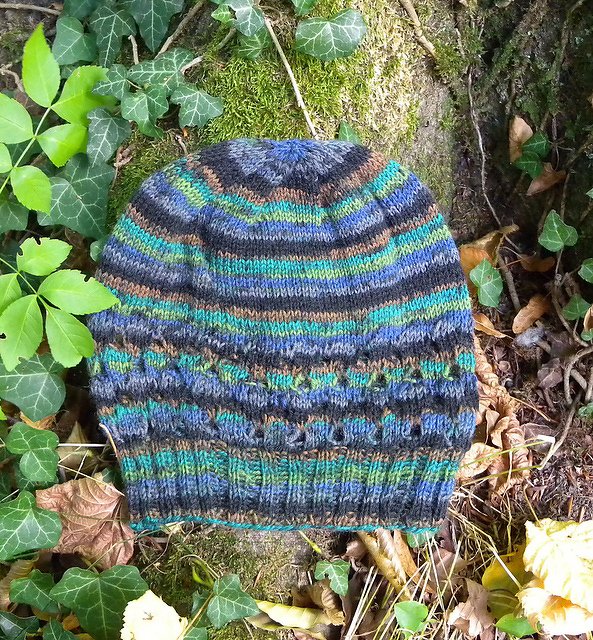 Striped knitted beanie in blue, green, brown and grey with textured stitch band, placed on moss and leaves.