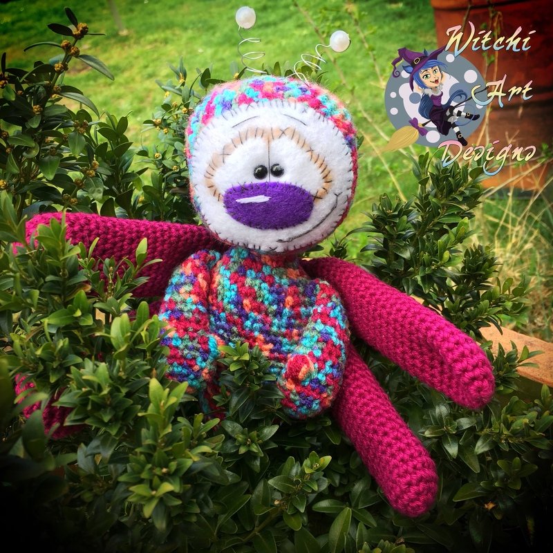 Buntes gehäkeltes Amigurumi mit spiralförmigen Antennen und pinken Armen zwischen grünen Blättern.