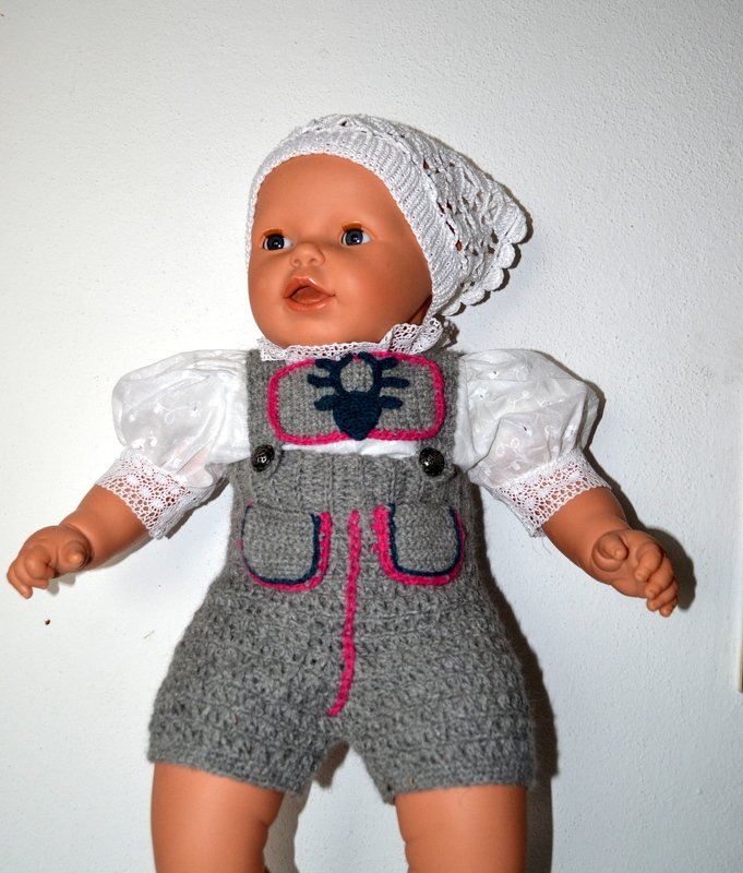 Gehäkelte kurze Baby-Latzhose in Grau mit Knöpfen, Taschen und rosa Ziernaht an Babypuppe
