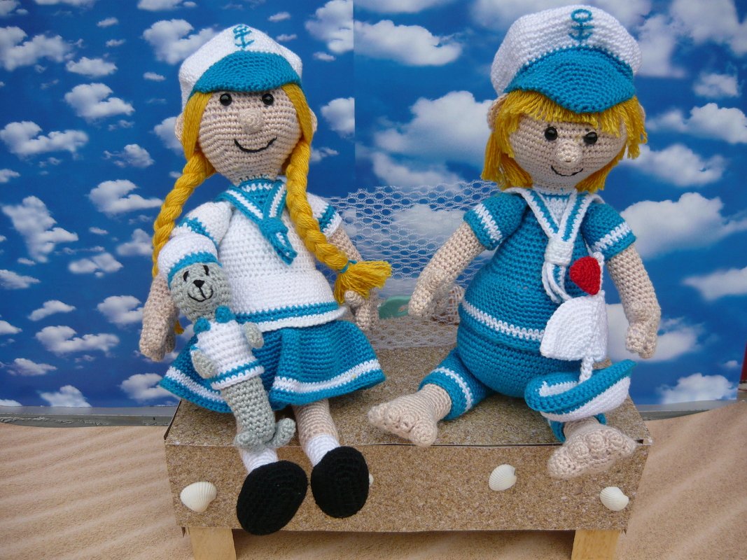 Zwei gehäkelte Amigurumi-Matrosenpuppen in blau-weißen Outfits, Mädchen mit Zöpfen und Robbe, Junge mit Segelboot vor wolkenblauem Hintergrund