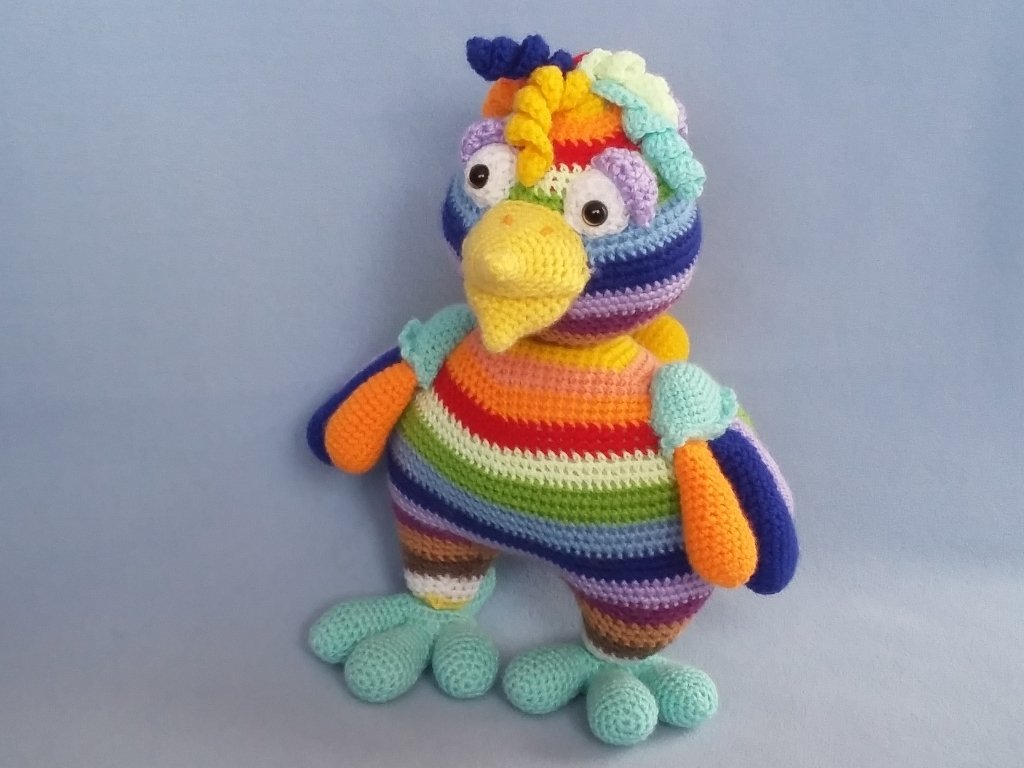 Gehäkelter Amigurumi-Vogel in Regenbogenstreifen mit gelbem Schnabel und hellblauen Füßen vor blauem Hintergrund.
