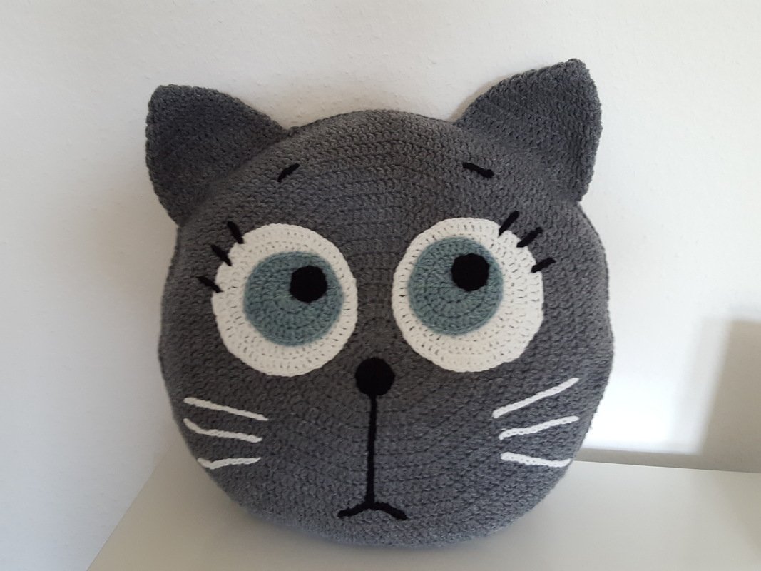 Gehäkeltes graues Katzenkopfkissen mit weißen Augenringen, hellblauer Iris, gestickter Nase und Schnurrhaaren.
