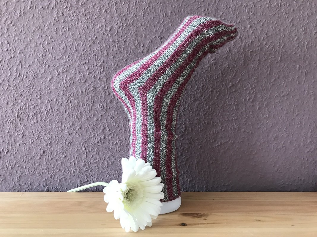 Gestreifte Stricksocke in Pink und Grau auf weißem Fußmodell mit weißer Blüte davor.