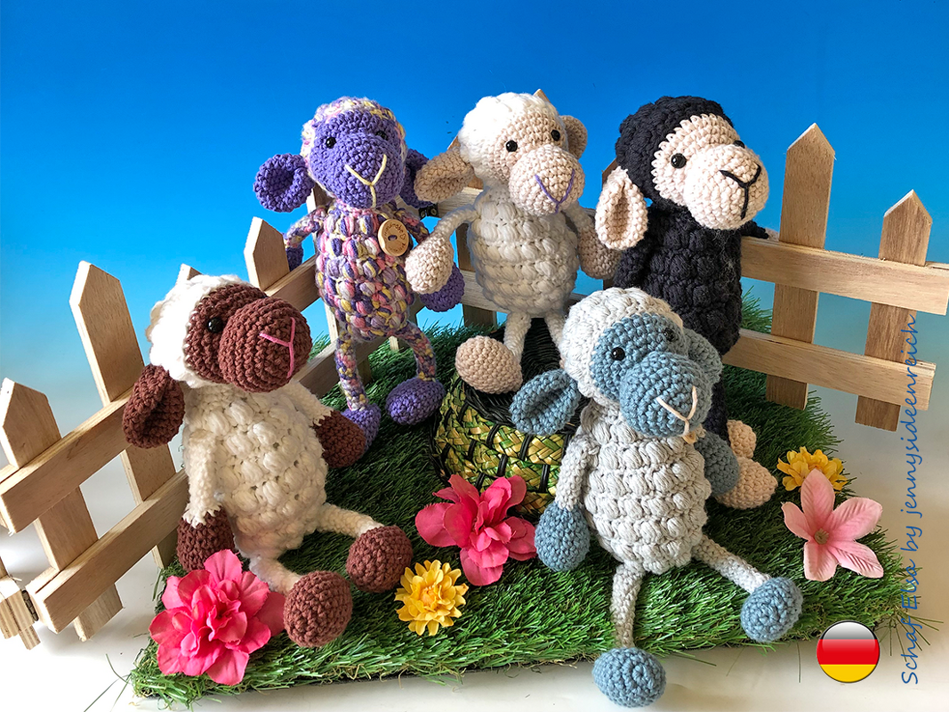 Fünf gehäkelte Amigurumi-Schafe in verschiedenen Farben auf Kunstgras mit Holzzaun und Blumen