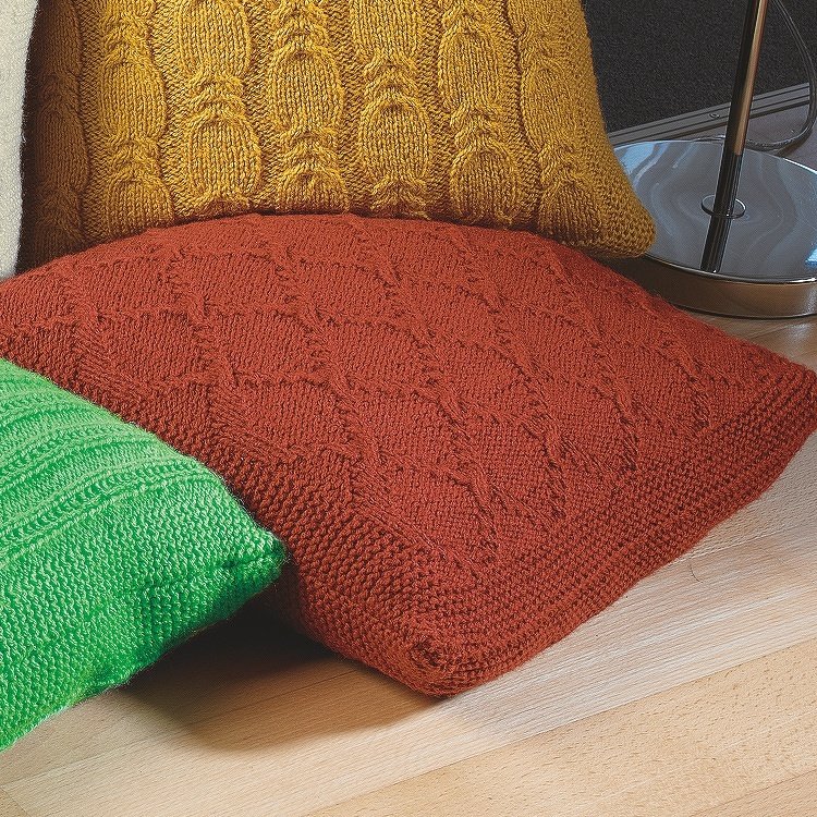 Gestrickte Kissen in Senf, Rost und Grün mit Zopf-, Rauten- und Rippenmustern, nebeneinander arrangiert