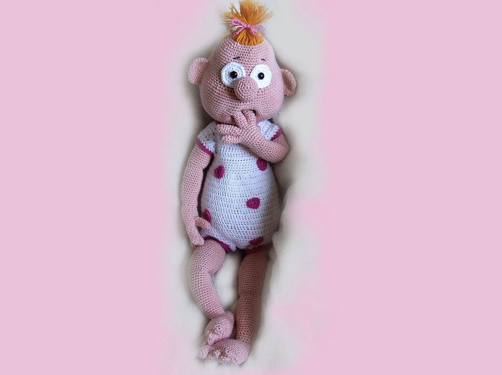 Rosa Amigurumi-Puppe mit weißem Strampler, pinken Punkten und orangefarbenem Haarschopf, mit großen, ausdrucksvollen Augen.