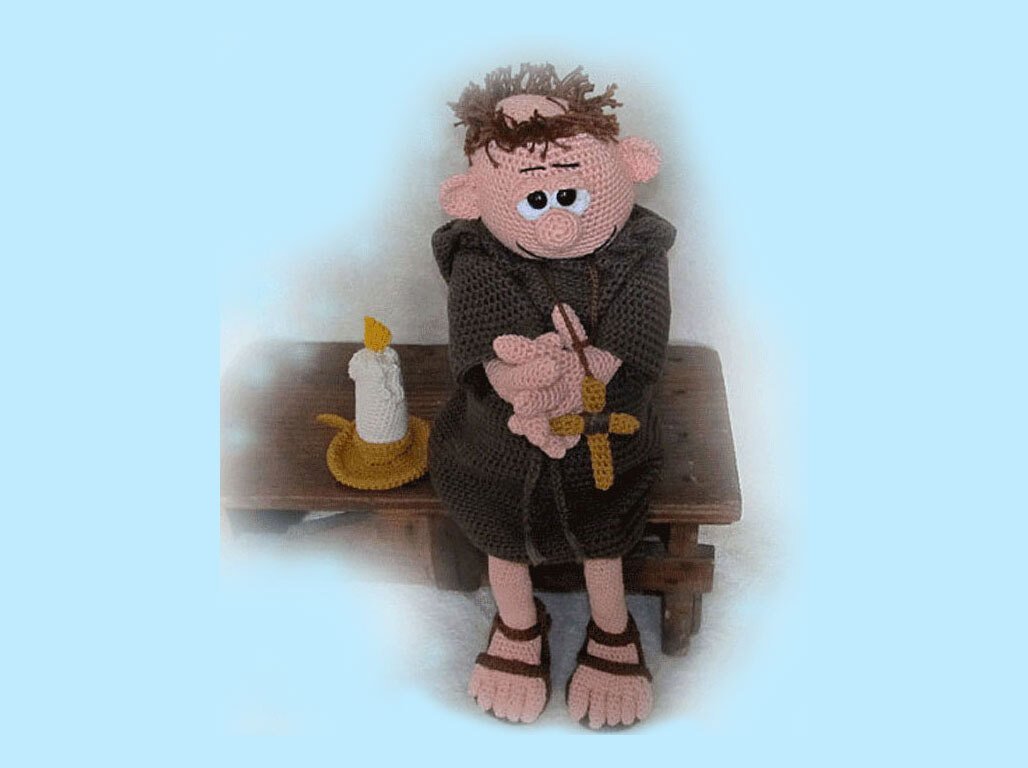 Amigurumi monk doll in brown habit holding a cross beside a crochet candle on a wooden bench
