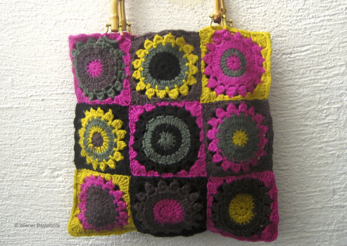 Gehäkelte Henkeltasche aus neun Granny Squares in Fuchsia, Gelb und Dunkelgrau mit Bambusgriffen vor heller Wand