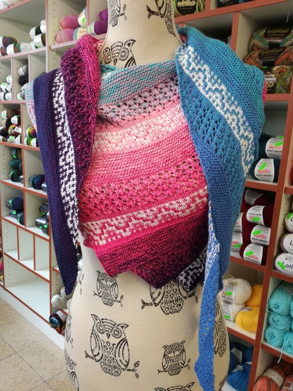 Gestrickter Drachenschwanz‑Schal mit Farbverlauf von Pink zu Blau auf Schneiderpuppe vor Garnregalen