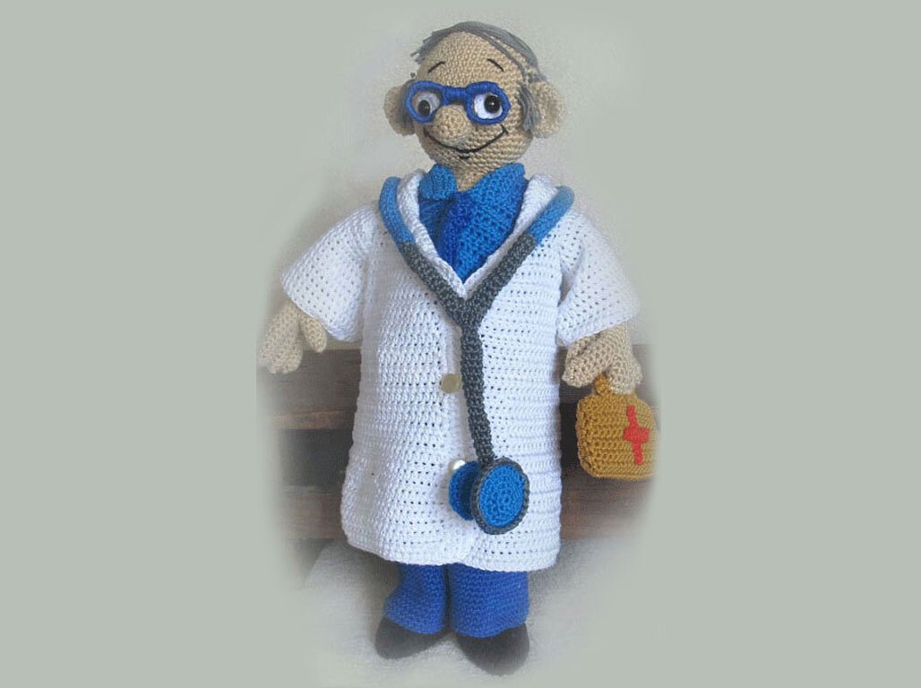 Amigurumi doctor doll wearing a white lab coat, blue glasses, a stethoscope, and holding a small brown first aid bag.