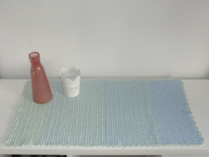 Handgehäkelter rechteckiger Tischläufer in Hellgrün bis Hellblau mit gewelltem Rand, daneben rosafarbene Flasche und weißes Windlicht.
