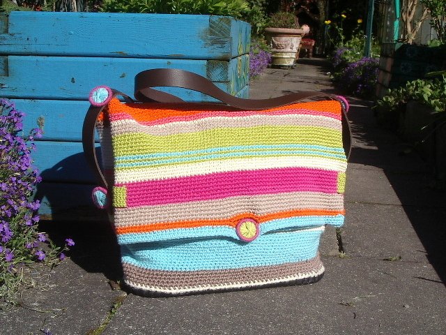 Gehäkelte Umhängetasche mit bunten horizontalen Streifen, Klappe, runden Knöpfen und braunem Schulterriemen vor blauem Holzkasten