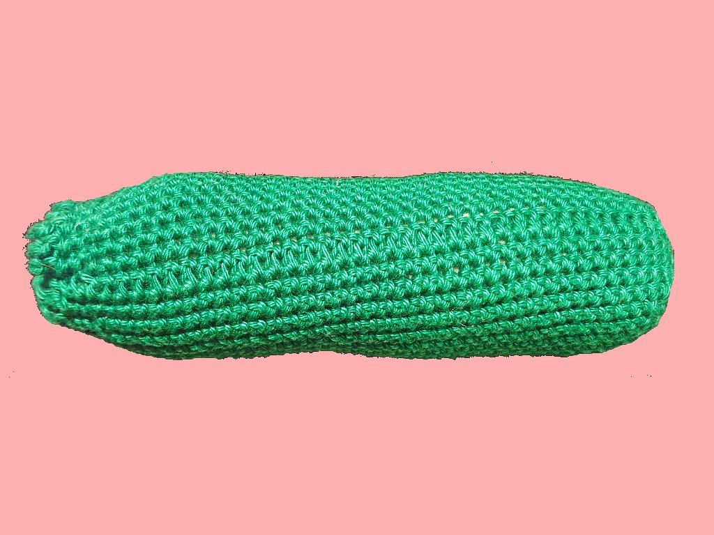 Längliche, grüne gehäkelte Gurke auf rosa Hintergrund.