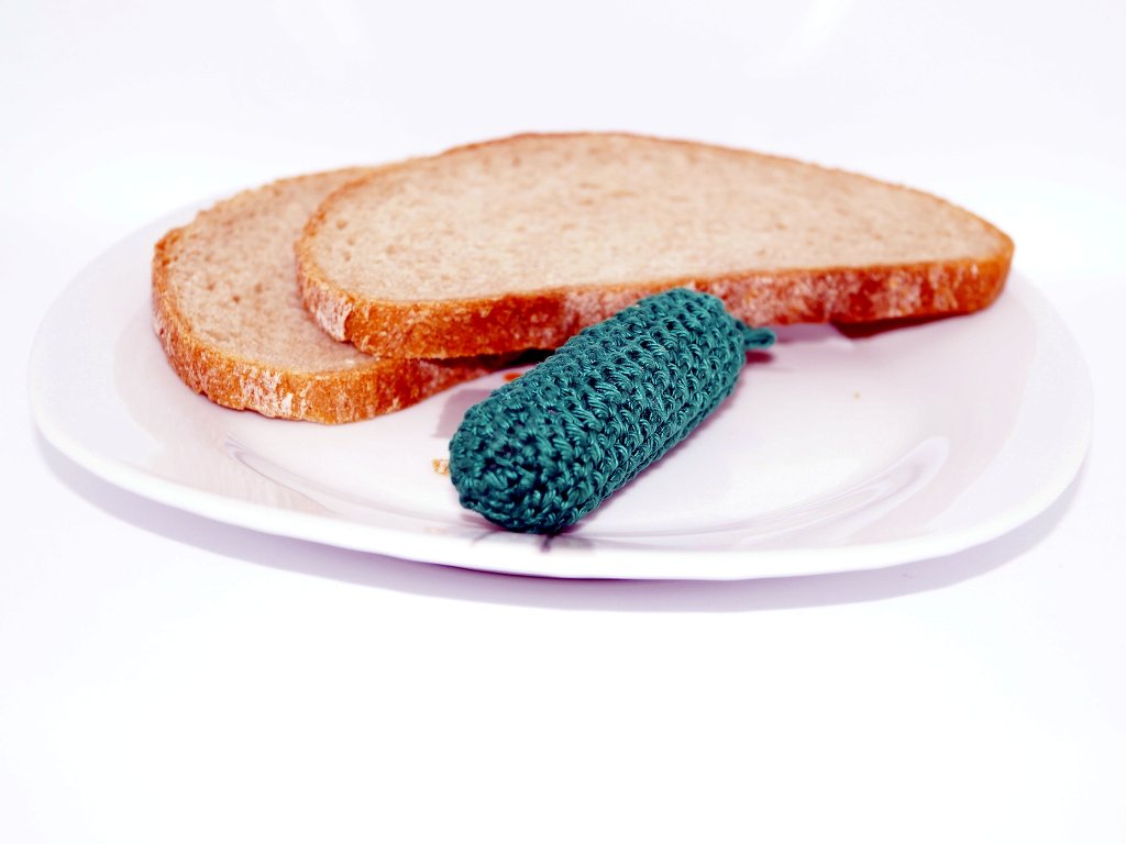 Gehäkelte grüne Gurke auf weißem Teller neben Brotscheiben.
