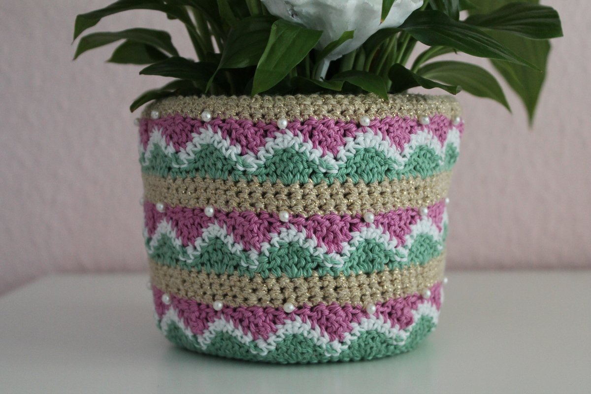 Gehäkelte Blumentopf‑Hülle mit Zickzackmuster in Rosa, Mint und Weiß, goldglänzende Reihen und aufgenähte Perlen