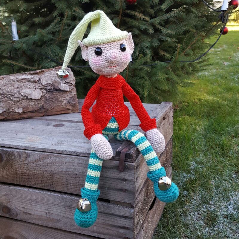 Gehäkelte Elfenpuppe mit roter Jacke, hellgrüner Zipfelmütze und gestreiften Beinen, sitzend auf einer Holzkiste vor einem Tannenbaum.