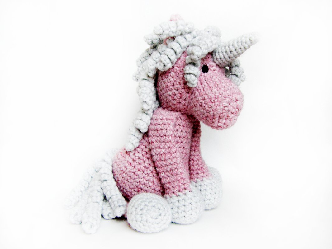 Gehäkeltes rosa Amigurumi‑Einhorn mit hellgrauer Mähne und Horn, sitzend