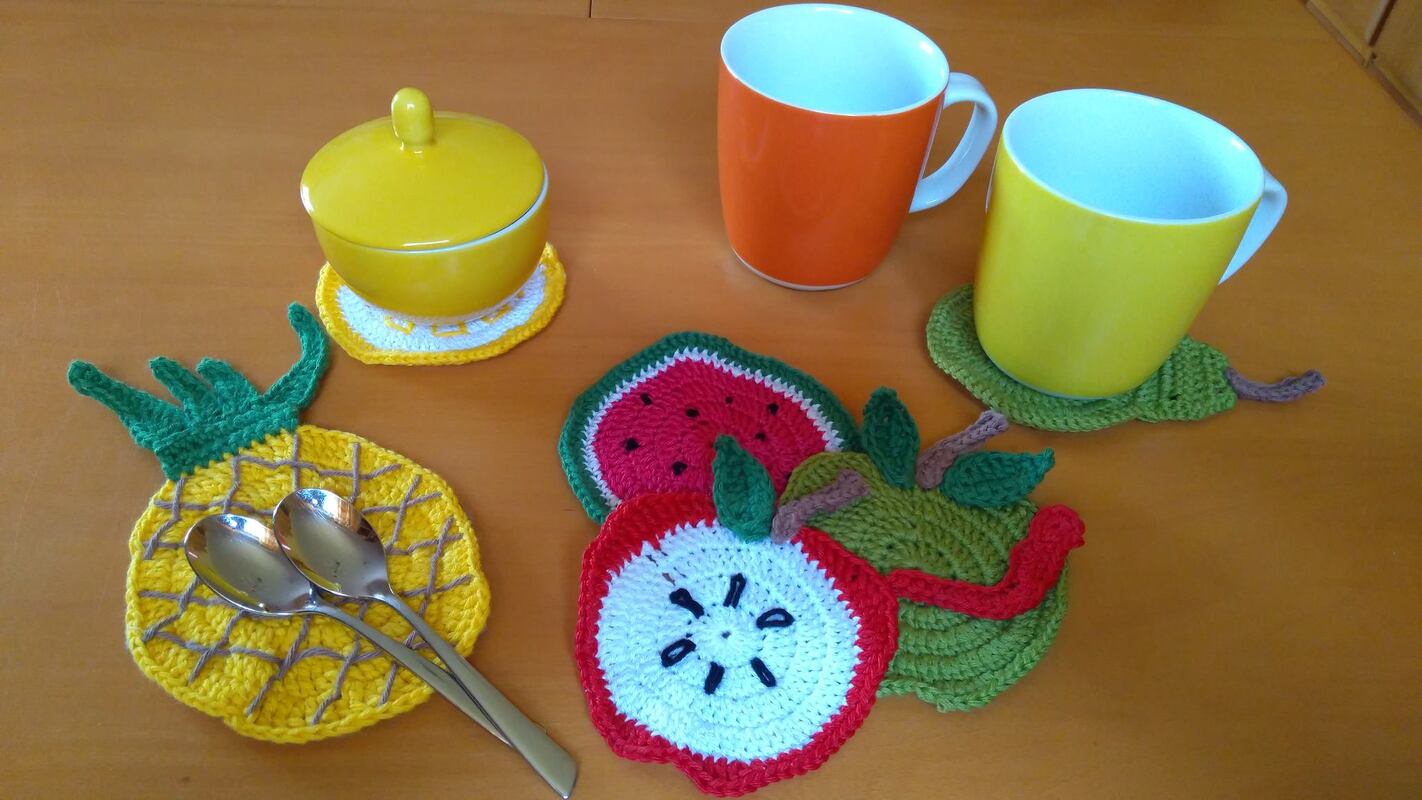 Set aus gehäkelten Frucht-Untersetzern: Ananas, Wassermelone, Apfelscheibe und grüner Apfel, die Tassen und ein Zuckergefäß auf einem Holztisch schmücken.