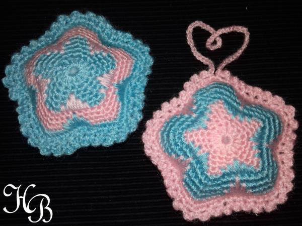 Zwei gehäkelte Sterne in Rosa und Hellblau auf dunklem Hintergrund, einer mit herzförmiger Schlaufe