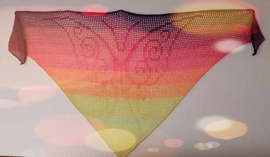 Gehäkeltes dreieckiges Tuch mit regenbogenfarbenem Farbverlauf und zentralem Schmetterlingsmotiv, an einer Wand aufgehängt