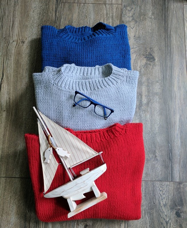 Drei gefaltete Strickpullover in Blau, Grau und Rot mit blauer Brille und Holzsegelboot auf Holzboden.