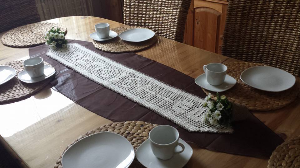 Weißer gehäkelter Tischläufer im Filetmuster auf dunklem Unterläufer, arrangiert auf einem Holztisch mit geflochtenen Platzsets und weißem Geschirr.
