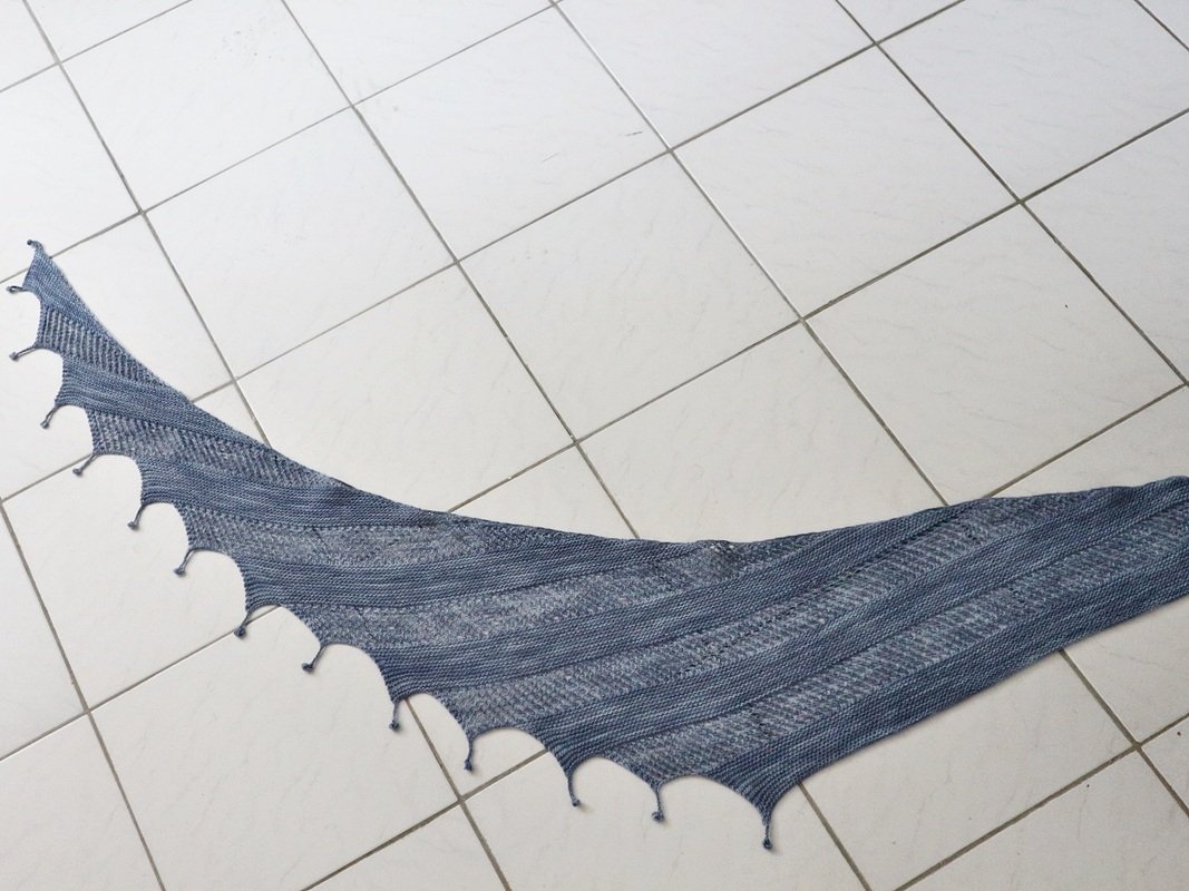 Gestrickter Drachenschwanz‑Schal in Blau‑Grau mit gestreiftem Muster und gezacktem Rand auf hellem Fliesenboden.