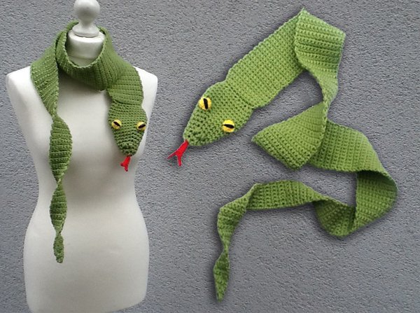 Grüner gehäkelter Schlangenschal mit gelben Augen und roter Zunge, präsentiert an einer Schaufensterpuppe und flach liegend.