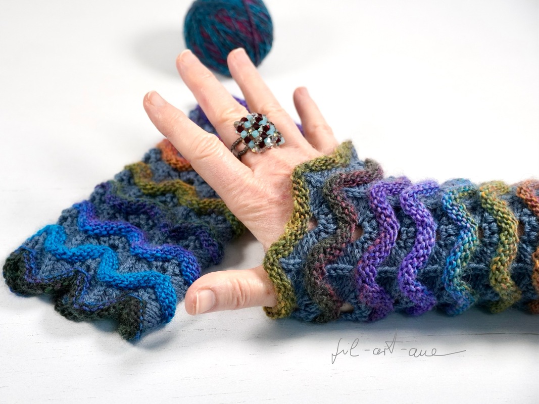 Hand wearing a colorful fingerless knitted glove with a wavy stitch; matching glove and yarn ball in the background.