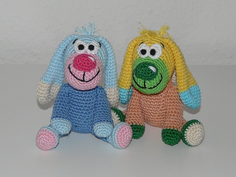 Zwei gehäkelte Amigurumi‑Hunde nebeneinander, einer hellblau‑rosa, einer gelb‑grün