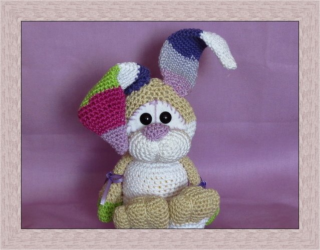 Sitzender Amigurumi-Hase in Beige und Weiß mit bunten Patchwork-Ohren vor violettem Hintergrund