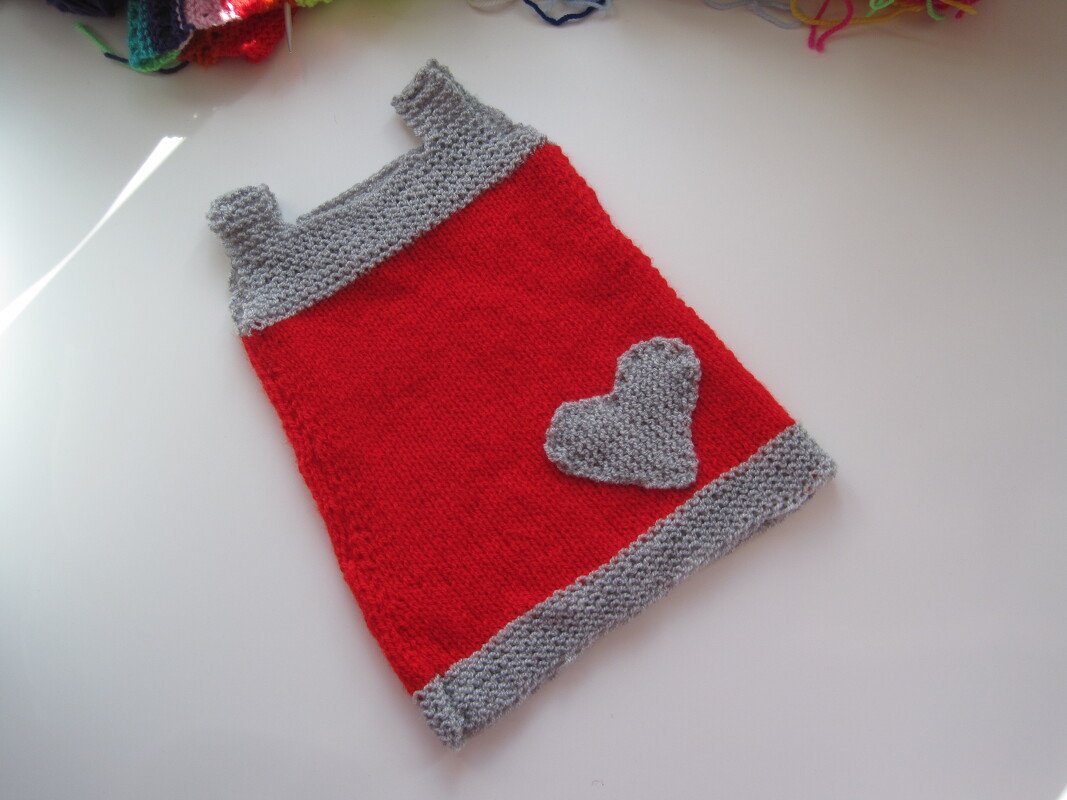 Rotes gestricktes Babykleid mit grauen Bündchen und grauem Herz auf weißem Hintergrund.