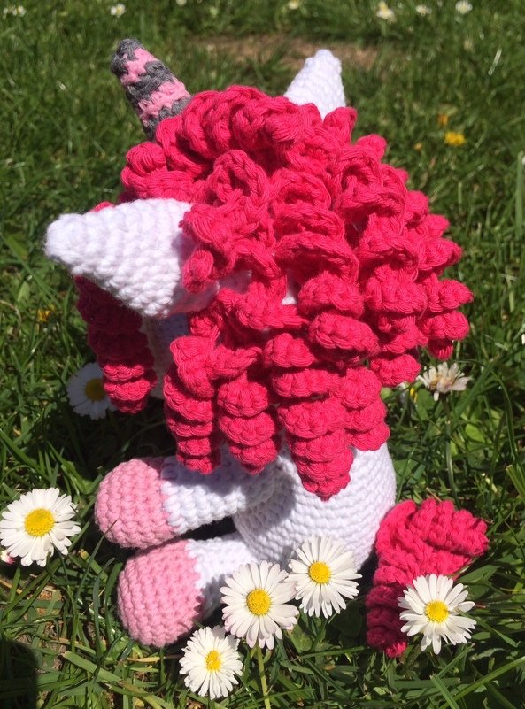 Gehäkeltes Einhorn-Amigurumi mit spiralförmigen Locken und gestreiftem Horn, sitzend auf Gras