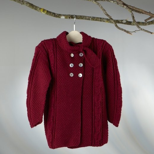 Dunkelrote gestrickte Kinderjacke mit sechs hellen Knöpfen und Schleife am Kragen, aufgehängt an einem Ast.